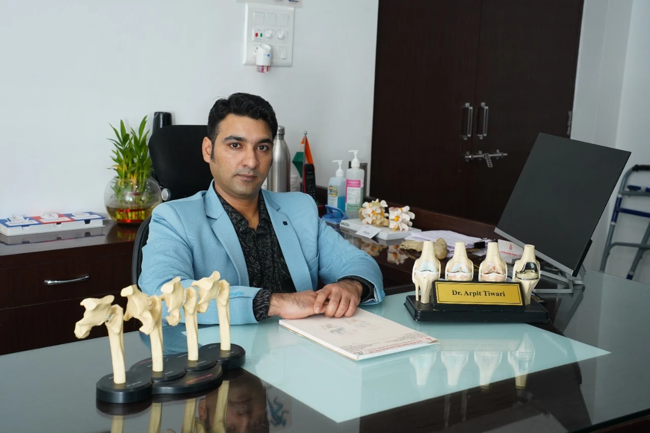 Orthopedic surgeon sitting in clinic with knee and joint models, representing joint replacement and orthopedic consultation.